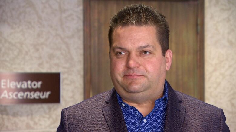 A man with brown hair wearing a suit is looking slightly off camera. He is standing in front of a wall with a sign that says 'elevator' behind him. 