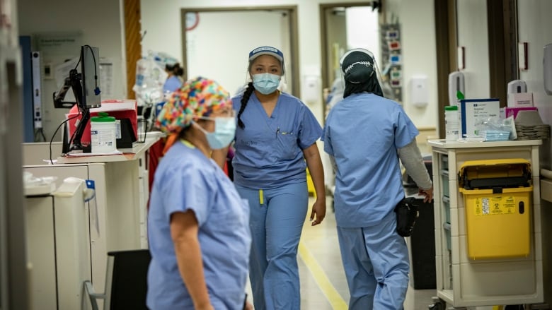 Three nurses, masked and wearing blue scrubs, walk in different directions through a hospital corridor. 