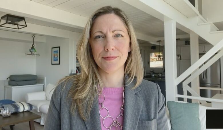 A woman with long hair wearing a rose-coloured blouse and a grey jacket speaks to the camera.