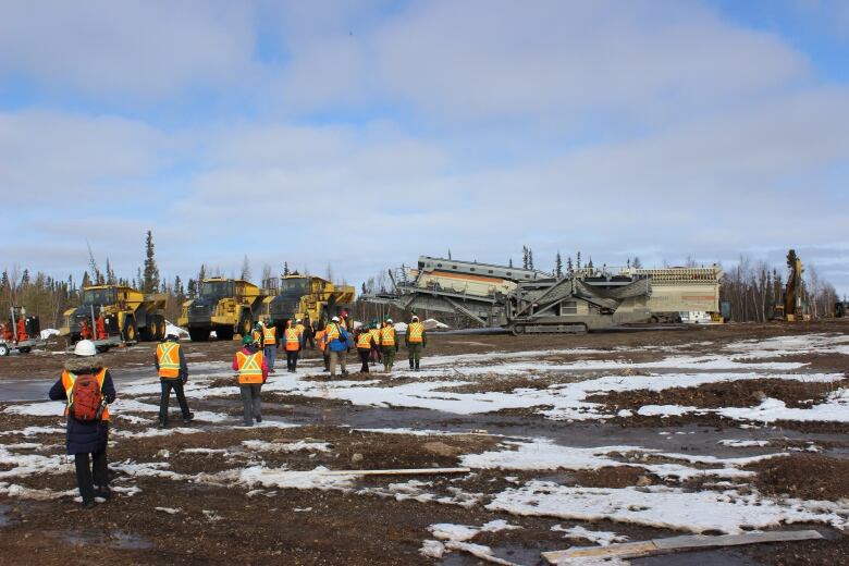 People in orange safety vests walking toward a row of heavy machinery. The ground is visible but snowy.