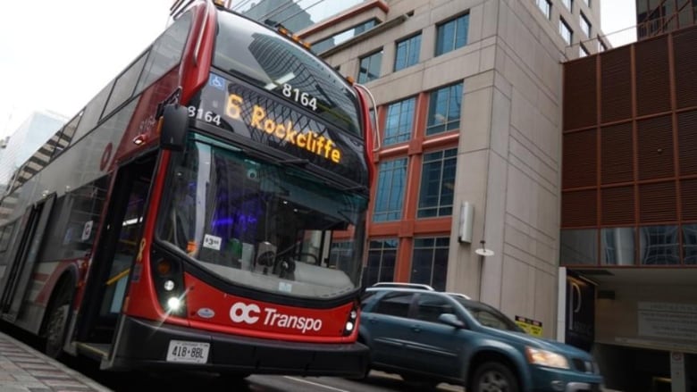 A double-decker red bus is parked on a downtown street while another car drives past.