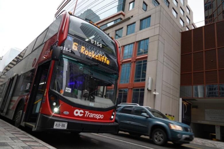 A double-decker red bus is parked on a downtown street while another car drives past.