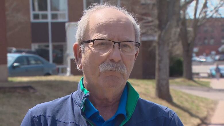 A white man with a moustache and glasses looks at the camera