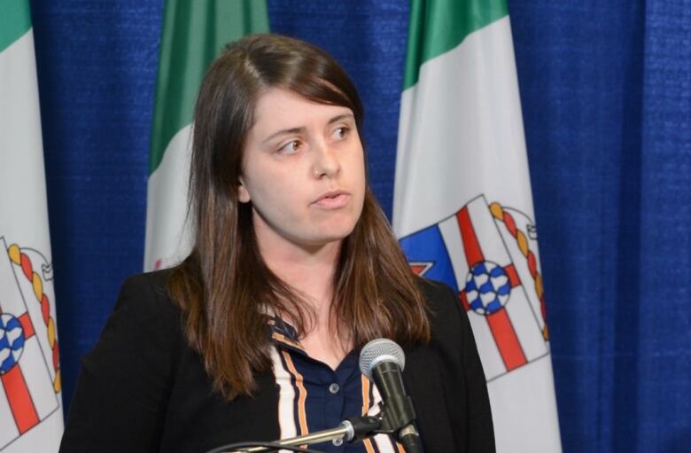 A woman stand at a microphone with flags and a curtain behind her.