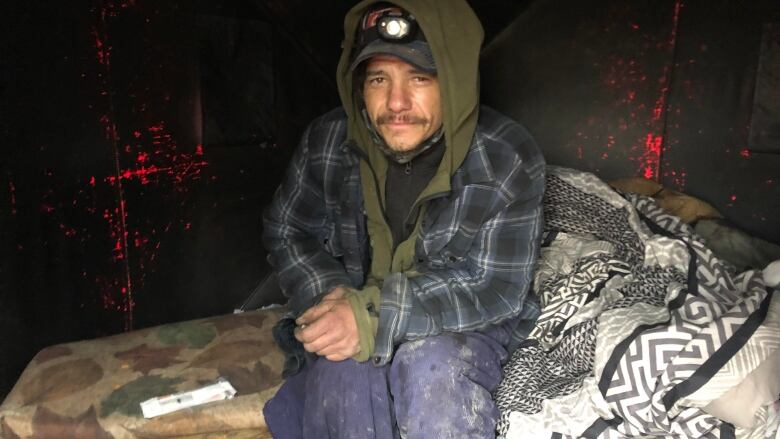 A photo of a man in many layers of clothes and a hood over his head, sitting on an older looking mattress. 