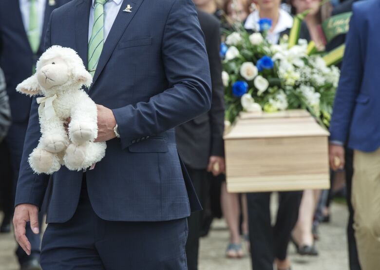 A man holds a stuffed animal. A casket is visible behind him. 