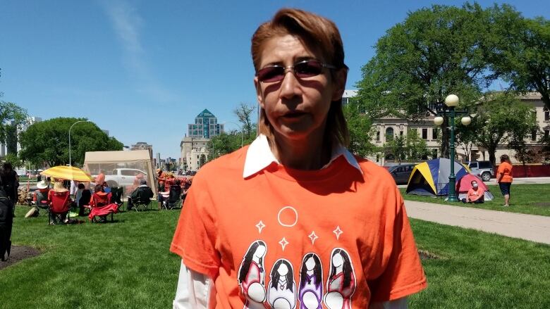 A woman with short red hair is pictured in an orange shirt that has an illustration of four women on it. Behind her are a number of tents and people.