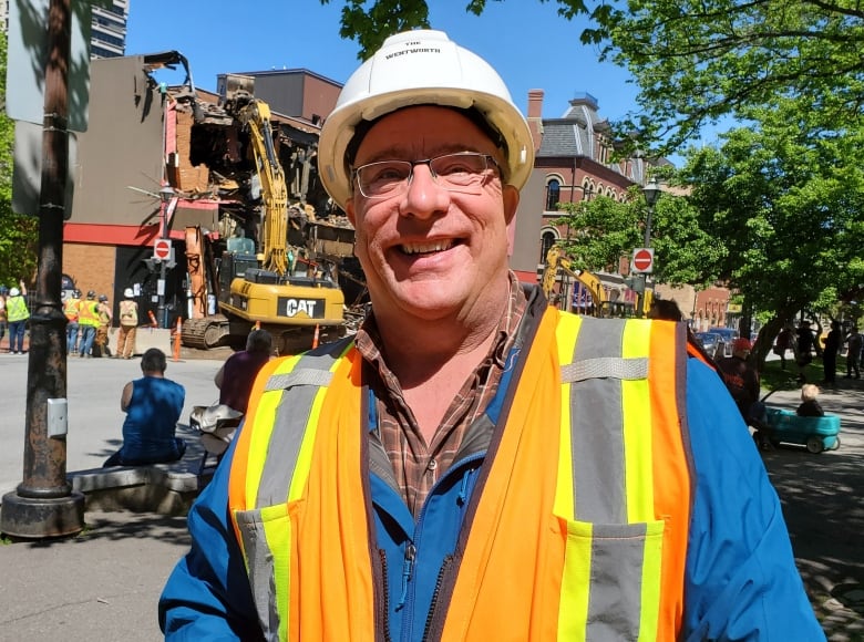 Man in hard hat and a safety vest in front of an excavator tearing down a building.