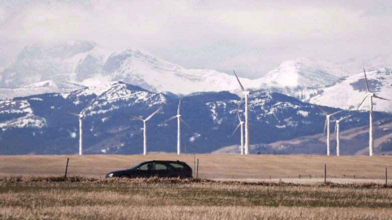 A row of wind turbines is set against a backdrop of mountains. A black car can be seen in the foreground. 