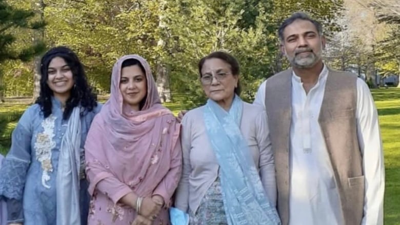 A family stands outside, looking to the camera. There are trees in the background.