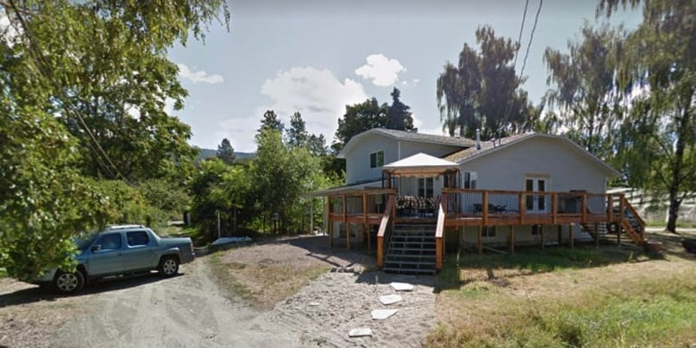 A home with two peaked roofs at the end of a dirt driveway has a car parked outside it.