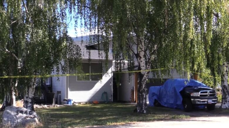 Pictured is a white house with a yard full of willow trees and a truck half covered by a blue tarp. 