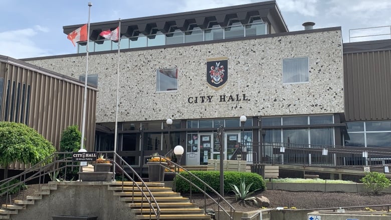 Brown stone steps lead up to a glass-fronted exterior below a white stone wall that says City Hall in black, topped by a coat of arms.