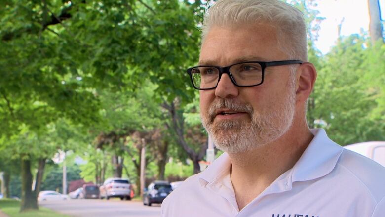 A white man with white hair is seen wearing black glasses, and a white polo shirt.