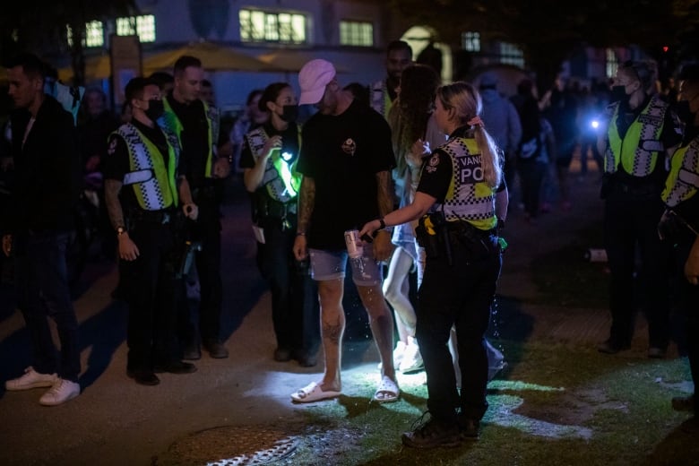 A police officer pours out a can of alcohol while looking at a man wearing a hat, as other police officers look on.