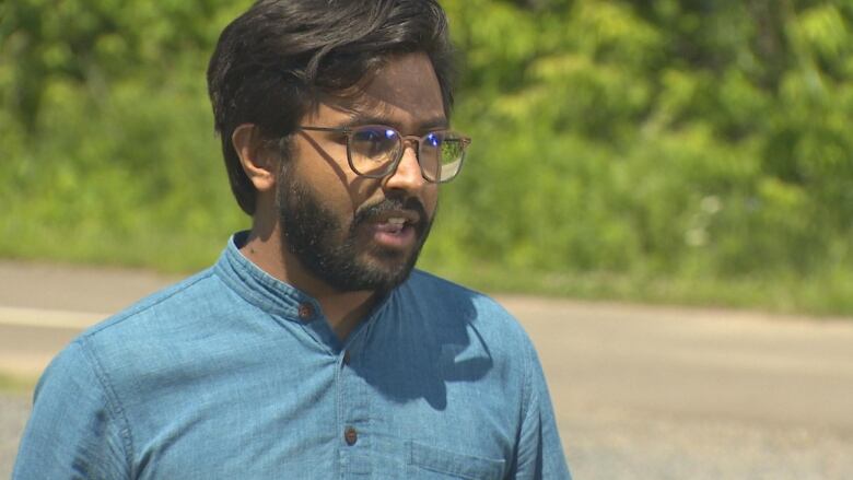 A man is a blue shirt stands outside speaking to a camera.