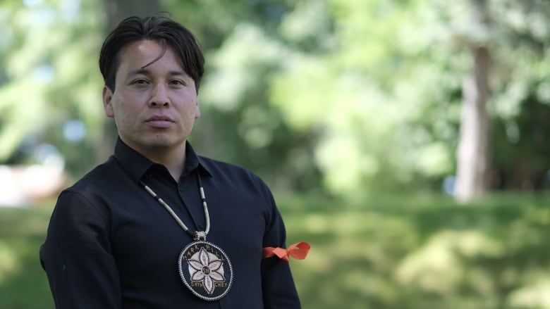 A man wearing a beaded medallion stands in a park.