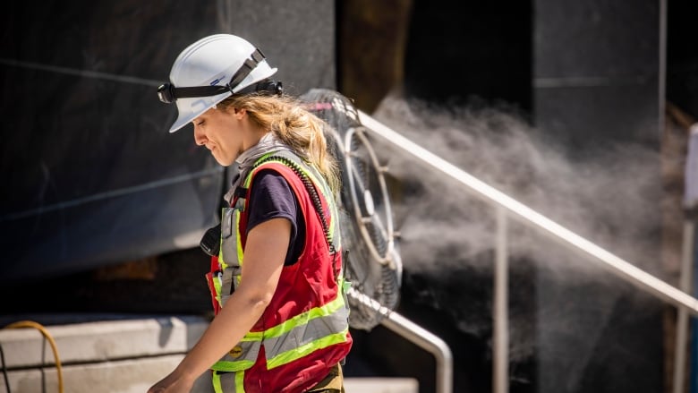 A female construction worker wearing a red safety vest and a white hard hat walks by a cooling fan on a job site.