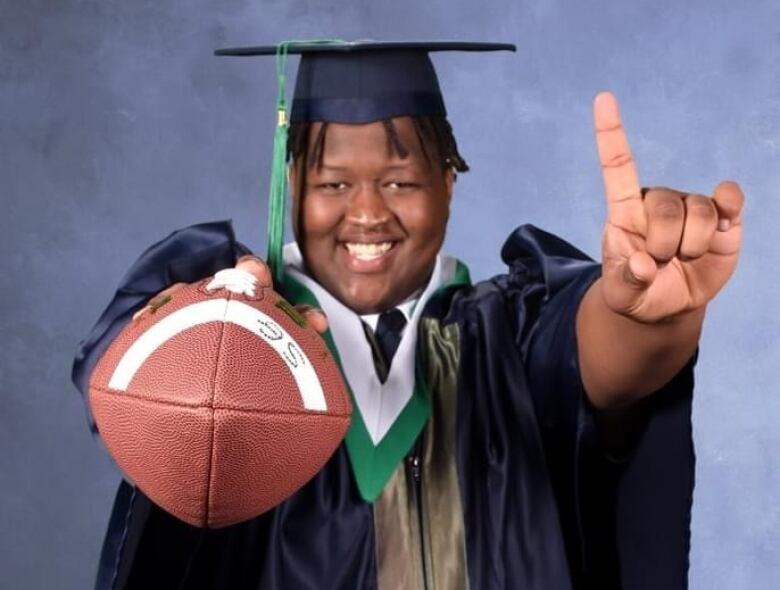 A man in a cap and gown holds a football and a forefinger up and smiles at the camera.