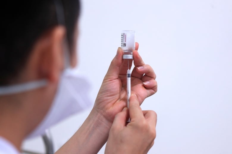 A person wearing a medical mask draws liquid from a vial with a syringe. Their face is visible from profile.