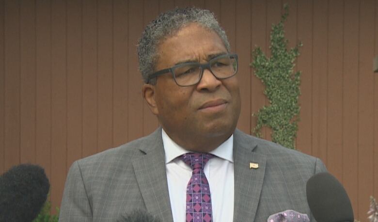 A Black man is seen wearing glasses and a grey suit. He is standing in front of several mics.