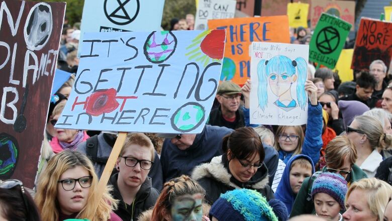 A large crowd stands with signs with climate-change related slogans. One says It's getting hot in here, another says there is no planet B. 