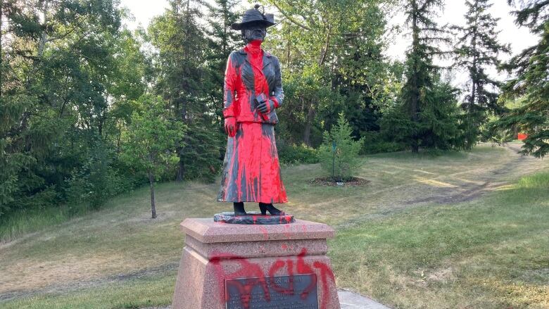 A statue of a woman in a park is defaced with red paint.