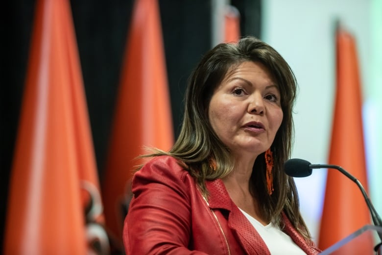 A woman in a red blazer sits at a table with orange flags behind.