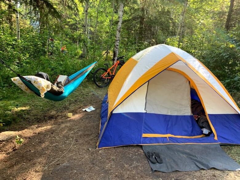 A blue tent with yellow trim sits in a wooded campsite. To the left of it, tied between two trees, is a teal hammock with a person inside reading a book. Their face is not visible.