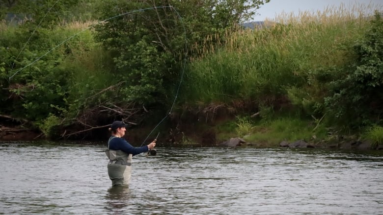 A woman stands in a river and casts a fly rod backwards while fishing.