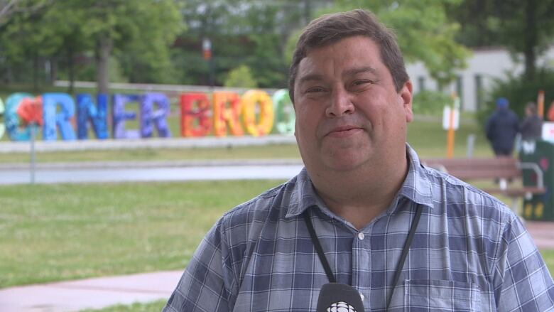 A man in a plaid shirt is shown outdoors in a grassy are with trees, and a large Corner Brook sign in the background.