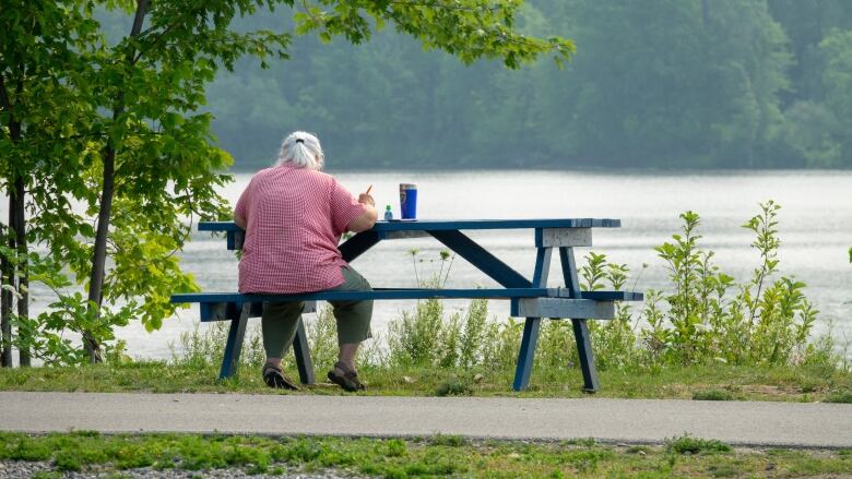 A woman sits outside at a green picnic table located next to a river.