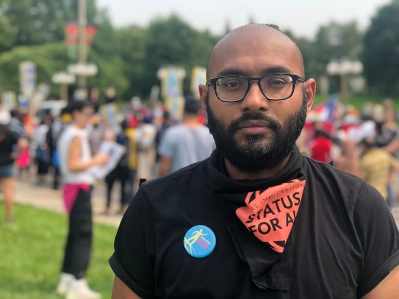 A man in glass and a scarf looks into a camera. A crowed of protesters can be seen in the background.