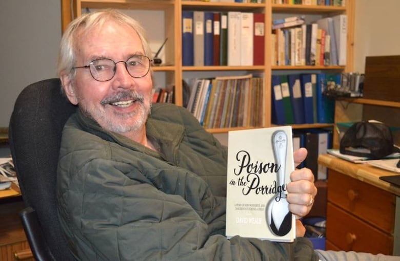 A smiling man with glasses and white hair holds up a book. 