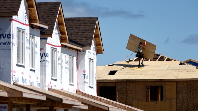 A man carries a piece of plywood across the roof of a home under construction