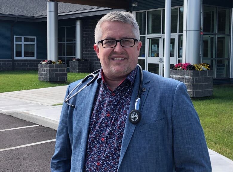 A person in a blue suit with a stethoscope around his neck stands in a parking lot in front of a building. 