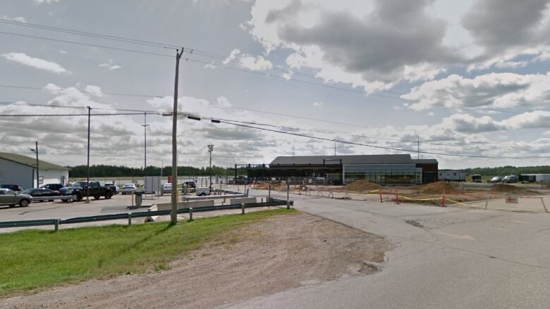 Wide pavement roads and electrical wires hang over a long, short building in the distance. The sky is blue with several grey clouds. 