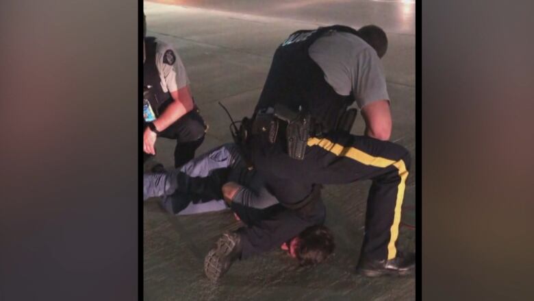 Two uniformed RCMP officers hold down a man lying on a sidewalk, with one of the officers' knees on the man's neck.