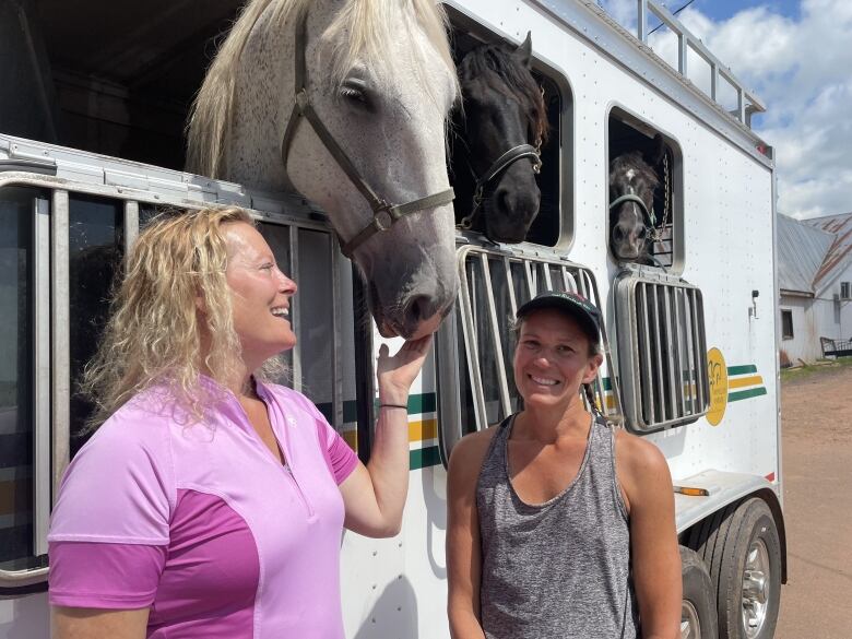 Two woman stand by horse trailer with horses leaning out. 