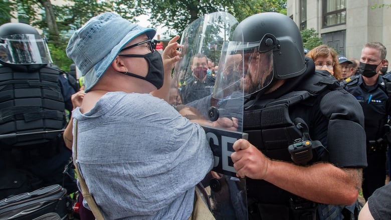 A person wearing a face mask faces off against a police officer in riot gear.