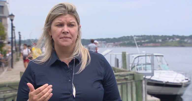 A woman with blonde hair in a blue jacket speaks in front of boats and water.