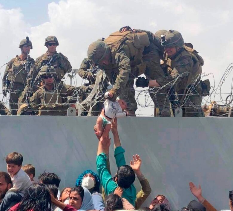 Troops in camoflage reach down over barbed wire to grab a baby being passed up to them