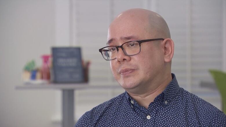 A bald man wearing glasses sits and talks to the camera.