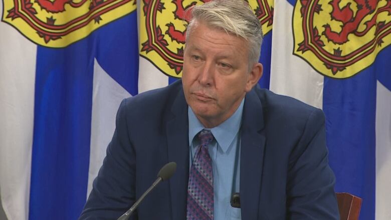 A man in a suit sits in front of a Nova Scotia flag. 