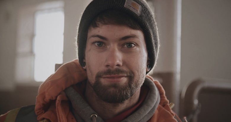 A portrait of a man with a grey hat, brown hair and a brown beard. He's wearing an orange sweatshirt.