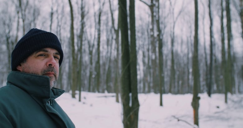 A man in a black toque and a green jacket stands outside in a forest in the winter. There is snow on the ground. 