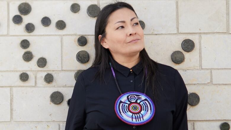 A woman wearing a black shirt and beaded necklace is standing in front of a brick wall and looking off to the side.