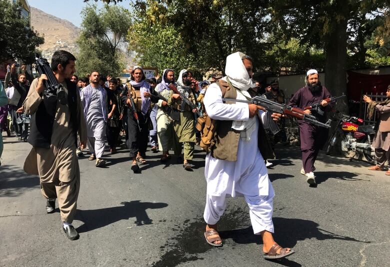 A group of men dressed in traditional Afghan clothes and armed with rifles walk down a crowded city street.