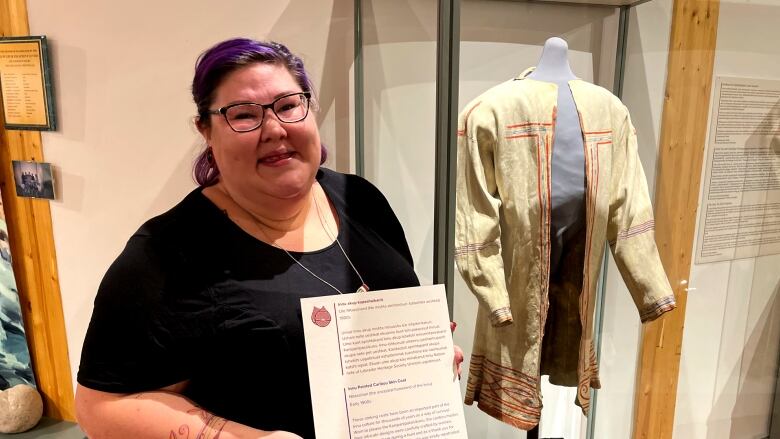 A woman holds a plaque for an Innu coat. A painted caribou hide coat is behind her.