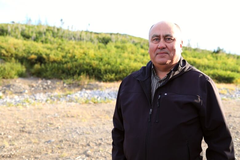 A man in a black jacket stands in front of a hill. 